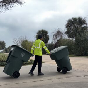 Person moving two large green trash bins on a driveway.