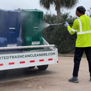Worker pressure washes trash bins on a truck in safety gear.