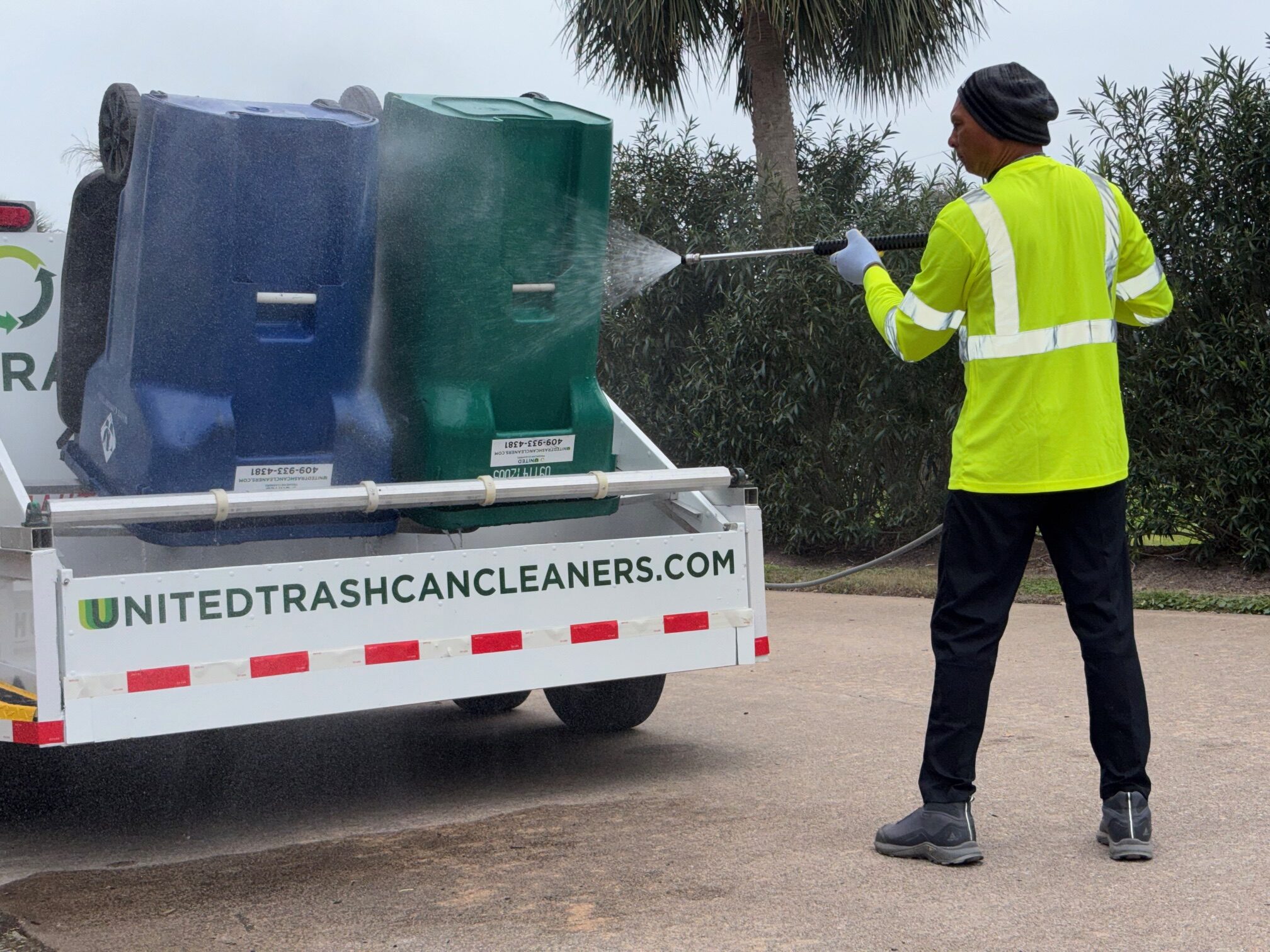 Worker pressure washes trash bins on a truck in safety gear.